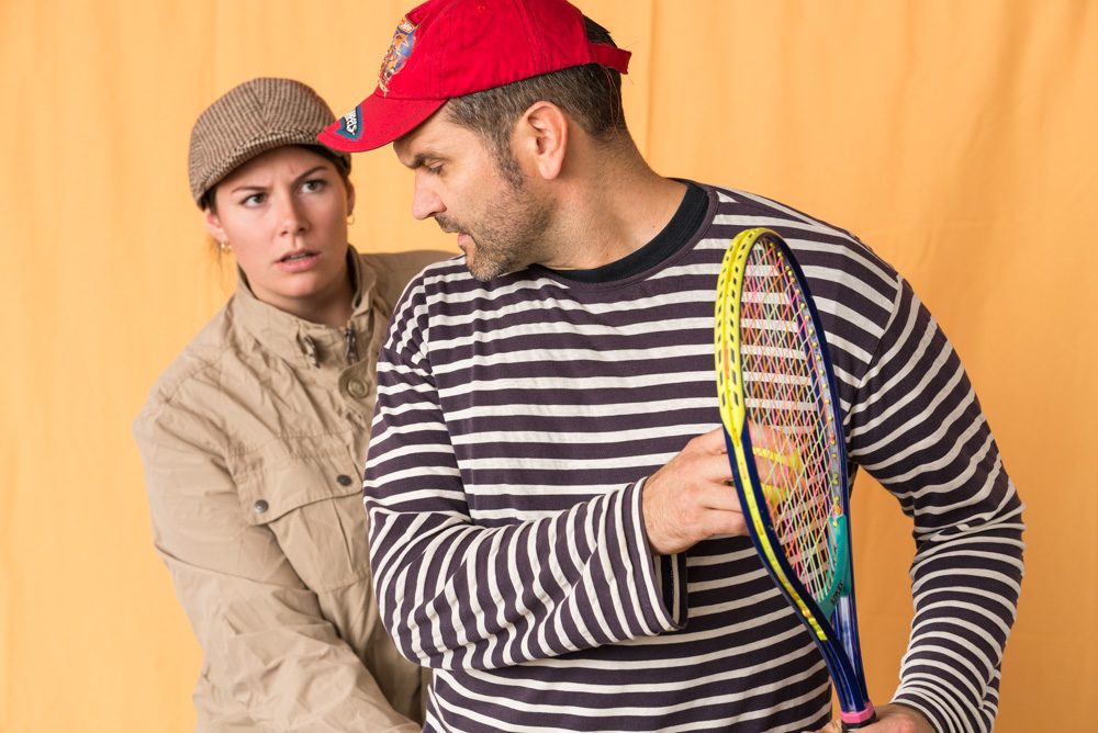 Theaterszene: ein Mann mit Tennisschläger und rotem Cap, dahinter eine Frau mit Schiebermütze